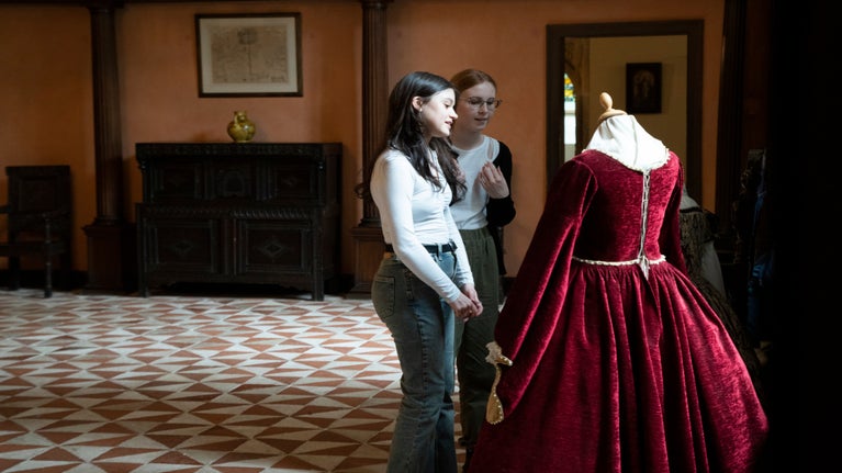 Two young women stand beside a rich red Tudor dress in the Great Hall of Buckland Abbey.